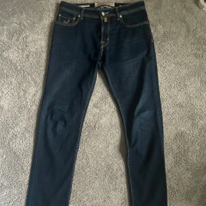 Mörkblå jeans från Jacob Cohen - Snygga mörkblå jeans från Jacob Cohen med kontrasterande sömmar och klassisk femficksdesign. De har en knappgylf och en stilren läderpatch med logotyp baktill. Perfekta för en stilren look. Skick 9,5/10, Endast använda två till tre gånger. 