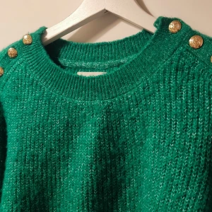 Grön stickad tröja från Holly & Whyte - Snygg grön stickad tröja från Holly & Whyte med dekorativa guldknappar på axlarna. Perfekt för att hålla sig varm och stilren under kyligare dagar. Tröjan har en klassisk rund halsringning och ribbade muddar.