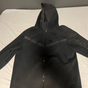 Svart hoodie från Nike - Säljer en svart hoodie från Nike med dragkedja och huva. Jackan har en praktisk ficka med dragkedja på ärmen och är perfekt för en sportig look. 