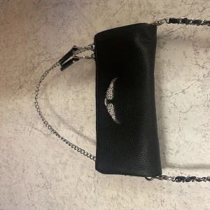 Zadig liknande väska i svart - Snygg svart axelväska i skinn med en cool kedjerem och ett glittrigt vingmotiv på framsidan. Perfekt för att ge din outfit en edgy touch. Väskan har en dragkedja för säker förvaring av dina saker.