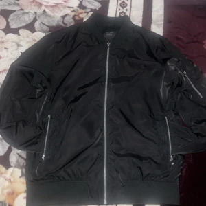 Svart bomberjacka med dragkedja - Snygg svart bomberjacka från Jack and Jones  med dragkedja framtill och på fickorna. Jackan har en klassisk bomberstil med ribbade muddar vid ärmslut och nederkant. Perfekt för en stilren look.