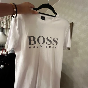 Vit t-shirt från Hugo Boss - Säljer en stilren vit t-shirt från Hugo Boss med deras ikoniska logga tryckt på framsidan. T-shirten har en klassisk passform och är tillverkad i mjukt bomullsmaterial. Perfekt för en avslappnad och trendig look.