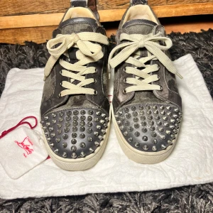 Grå sneakers med nitar från Christian Louboutin - Snygga grå sneakers från Christian Louboutin med nitar på tån och klassisk röd sula. Skorna har snörning och är tillverkade i mocka med en cool camouflage-look. Perfekta för att ge din stil en edgy touch. Dustbags ingår.