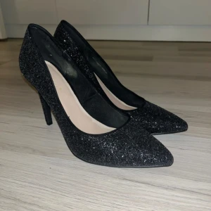 Svarta glittriga pumps - Snygga svarta glittriga pumps med spetsig tå och klack. Perfekta för en kväll ute eller en festlig tillställning. De har en elegant design som ger en lyxig känsla.