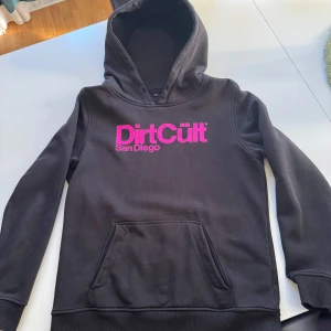 Svart hoodie från DirtCült - Säljer en svart hoodie från DirtCült med rosa tryck 'San Diego' på framsidan. Tröjan har en klassisk känguruficka och en bekväm huva. Perfekt för en avslappnad stil.