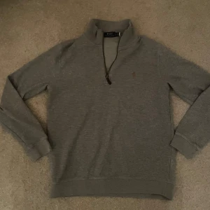 Grå tröja från Polo Ralph Lauren - Säljer en stilren grå tröja från Polo Ralph Lauren med halvsip vid halsen. Tröjan har långa ärmar och en liten broderad logotyp på bröstet. Perfekt för en avslappnad och snygg look.