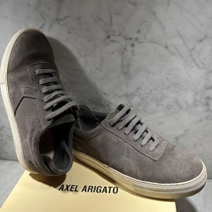Grå fräscha sneakers från Axel Arigato - Snygga grå sneakers från Axel Arigato i mocka med klassisk snörning. Skorna har en stilren design med en vit sula. Box och påse ingår. Nypris: 2449 kr