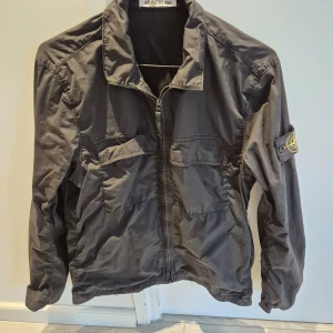 Svart jacka från Stone Island - Säljer en stilren svart jacka från Stone Island med dragkedja och klassisk krage. Jackan har en ikonisk logga på ärmen och är perfekt för en cool och avslappnad stil. Passar bra till både vardag och speciella tillfällen.