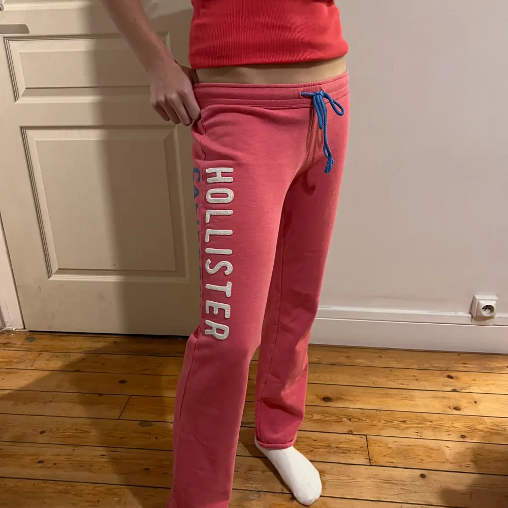 Snygga rosa mjukisbyxor från Hollister med texten 'Hollister California' längs benet. Byxorna har en blå snörning i midjan för justerbar passform. Perfekta för en avslappnad stil.. Farkut & Housut.