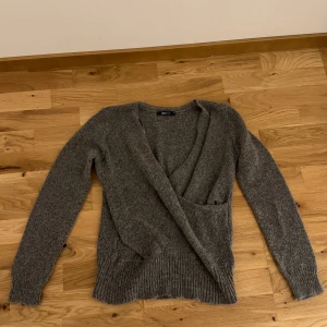 Grå omlottkofta från Gina tricot  - Säljer en mysig grå omlottkofta. Den har långa ärmar och ribbade muddar vid ärmslut och nederkant. Perfekt för kyliga dagar!