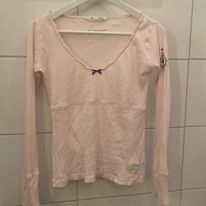 Ljusrosa ribbad tröja från Odd Molly - Säljer en ljusrosa ribbad tröja från Odd Molly med en söt rosett framtill och ett kompassmotiv på ärmen. Tröjan har en u-ringad design med spetsdetaljer vid halsen och långa ärmar.