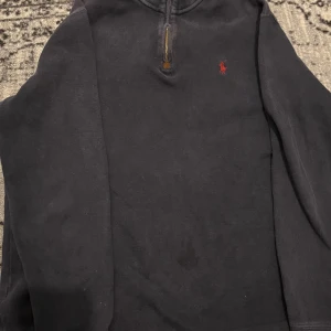 Mörkblå tröja från Ralph Lauren - Säljer en stilren mörkblå tröja från Ralph Lauren med en liten röd broderad logotyp på bröstet. Tröjan har en dragkedja vid halsen och långa ärmar. Perfekt för en avslappnad look. En mindre fläck på magen.