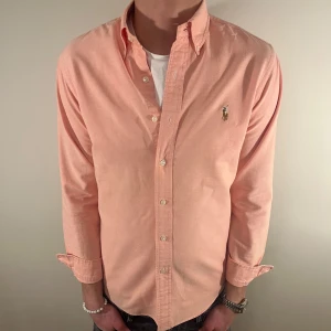 Rosa/orange Ralph Lauren skjorta - Nu säljer vi ännu en fet polo skjorta som passar till alla tillställningar! Skjortan har ett litet hål långst ned vid knap🔥parna(bild 3) därför sänker vi priset och skjortan är annars imycket bra skick!🤙Du ska kunna uttrycka stil utan att det kostar för mycket!🕺🍾💼🤩  Storlek👔: M Nypris💰: ca 1499 kr