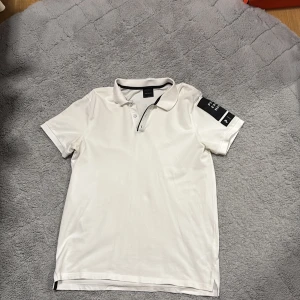 Vit pikétröja från Peak Performance - Säljer en stilren vit pikétröja från Peak Performance. Tröjan har korta ärmar och en klassisk krage med knappar. På ärmen finns en svart detalj med märkeslogga. Perfekt för en sportig och avslappnad look.