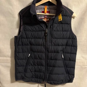 Parajumpers väst - Snygg svart dunväst från Parajumpers med dragkedja framtill och praktiska fickor. Perfekt för lager-på-lager under kyliga dagar. Den har en stilren design med en liten gul detalj vid kragen.