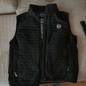 Svart quiltad väst från Colmar - Säljer en stilren svart quiltad väst från Colmar. Västen har dragkedja framtill och två praktiska fickor med dragkedjor. Perfekt för lager-på-lager under kyligare dagar. Märkeslogga på bröstet ger en sportig touch.