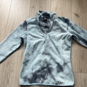 Blå fleece tröja från The North Face - Säljer en mjuk och bekväm blå fleece tröja från The North Face. Tröjan har en halv dragkedja framtill och ett subtilt mönster i mörkare nyanser. Perfekt för kyliga dagar och utomhusaktiviteter.