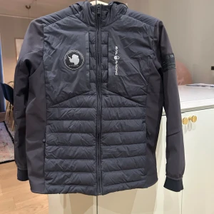 Svart jacka från Sail Racing - Snygg svart jacka från Sail Racing med dragkedja och huva. Jackan har ett stilrent märke på bröstet och är perfekt för kyligare dagar. Den quiltade designen ger extra värme och stil.