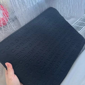 Svart datorväska från Marc Jacobs - Snygg svart datorväska från Marc Jacobs med präglad textdesign. Perfekt för att bära din laptop med stil. Enkel och elegant design som passar till alla tillfällen.