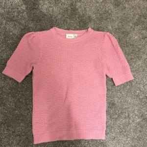 Rosa stickad tröja från Vila - Säljer en söt rosa stickad tröja från Vila. Tröjan har korta ärmar och en rund halsringning. Perfekt för en mysig och stilren look. 💖