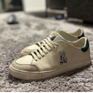 Arigato  - Snygga beige sneakers från Axel Arigato med en unik tecknad detalj på sidan. Skorna har grön häl och klassisk snörning. Perfekta för en stilren look.