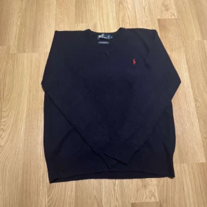 Mörkblå ulltröja från Polo Ralph Lauren - Säljer en stilren mörkblå tröja från Polo Ralph Lauren i 100% lammull. Tröjan har en klassisk v-ringning och det ikoniska röda logotypbroderiet på bröstet. Perfekt för kyligare dagar. Hör av dig vid intresse!