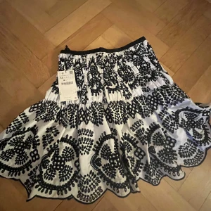 Vit och svart kjol med broderi - Charmig vit kjol med svart broderat mönster. Kjolen har en elastisk midja för bekväm passform och ett vackert, detaljerat broderi som ger en unik touch. Aldrig använd! Där med lappen kvar