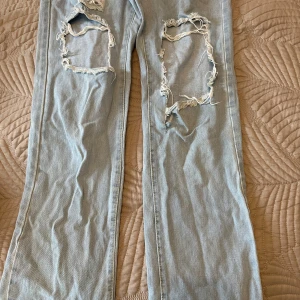 Ljusa jeans med slitningar - Säljer ett par ljusa jeans med stora slitningar på knäna. De har en straight passform och är perfekta för en avslappnad stil. Jeansen har en klassisk design med fem fickor och knappgylf.