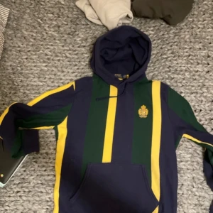 Randig hoodie från Ralph Lauren - Säljer en snygg hoodie från Ralph Lauren i blått, grönt och gult. Den har ett broderat emblem på bröstet och en klassisk huva. Perfekt för en avslappnad stil med en touch av elegans. Har en liten skarv på luvan men den syns inte typ 