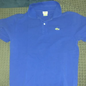 Blå pikétröja från Lacoste - Snygg blå pikétröja från Lacoste med klassisk krage och knappdetalj framtill. Den har korta ärmar och det ikoniska krokodilmärket broderat på bröstet. Perfekt för en stilren och avslappnad look.