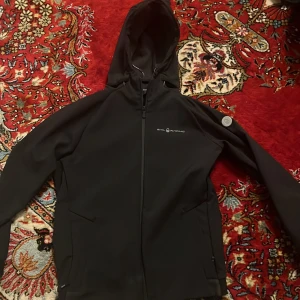 Svart softshelljacka från Sail Racing - Säljer en svart softshelljacka från Sail Racing med huva och dragkedja. Jackan har en stilren design med logotyp på bröstet och är tillverkad i polyester och spandex för komfort och rörelsefrihet. Helt ny, använd ungefär 2-3 gånger. Köpt för 1700 kr!