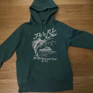 Grön hoodie från Polo Ralph Lauren - Säljer en grön hoodie från Polo Ralph Lauren med tryck på framsidan som visar en palm och texten 'Key West'. Hoodien har en justerbar huva med dragsko och är perfekt för en avslappnad stil. Den ör storlek M och jag är 185 och den sitter suveränt. (Pris kan diskuteras)