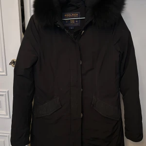 Svart parkajacka från Woolrich - Snygg svart parkajacka från Woolrich med pälskantad huva. Jackan har långa ärmar och knäppning framtill. Perfekt för kyliga dagar med sin stilrena design och praktiska fickor. Storlek S 