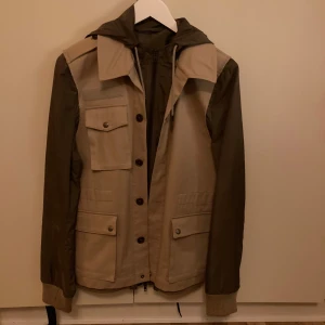 Beige och brun jacka från Maison Margiela - Snygg jacka från Maison Margiela i beige och brunt. Jackan har en huva och flera fickor med knappar framtill. Den är tillverkad i Italien och har en dragkedja samt knappar för stängning. Perfekt för en stilren look. Nästan oanvänd, gick för omkring 20 000 när den släpptes