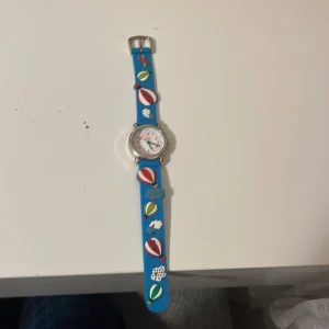 Blå barnklocka med luftballongmotiv - Charmig barnklocka med blått armband dekorerat med luftballonger och moln. Urtavlan har tydliga röda siffror och gröna visare, vilket gör det enkelt att läsa av tiden. Perfekt för barn som älskar färgglada och lekfulla accessoarer.