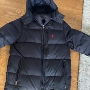 Mörk blå dunjacka från Ralph Lauren - Säljer en svart dunjacka från Ralph Lauren med huva och dragkedja. Jackan har en broderad logotyp på bröstet och flera praktiska fickor med dragkedjor. Perfekt för kyliga dagar.