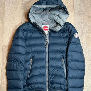 Mörkblå dunjacka från Colmar - Snygg mörkblå dunjacka från Colmar med huva och dragkedja framtill. Jackan har en quiltad design och två praktiska fickor med dragkedjor. Perfekt för kyliga dagar. Bara lite slitet på märket men de e lugnt 