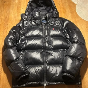 Svart pufferjacka från Ralph Lauren - Säljer en snygg svart pufferjacka i storlek XL från Ralph Lauren. Jackan har en quiltad design med dragkedja och huva, perfekt för kyliga dagar. Den är långärmad och har en liten röd logga på bröstet. Perfekt för att hålla värmen med stil!