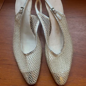 Glittriga guldklackar med nätmönster - Snygga klackskor i glittrigt guld med ett unikt nätmönster. Skorna har en elegant spetsig tå och en smal rem runt hälen. Perfekta för att ge en glamorös touch till din outfit.
