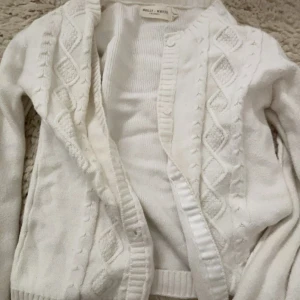Vit stickad kofta från Holly & White - Säljer en stilren vit stickad kofta från Holly & White. Koftan har ett klassiskt flätmönster och ribbade kanter. Perfekt för kyliga dagar och att ha som ett extra lager.