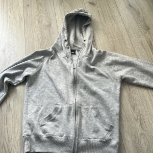 Grå hoodie från Cubus - Säljer en stilren grå hoodie från Cubus i storlek M. Den har en dragkedja framtill och en praktisk känguruficka. Perfekt för en avslappnad look. Huvan är mjuk och bekväm.