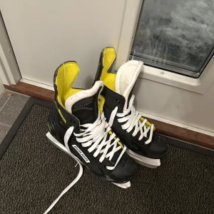 Svarta skridskor från Bauer - Säljer ett par svarta skridskor från Bauer med vita snören och gul insida. Perfekta för isaktiviteter och har en sportig design.