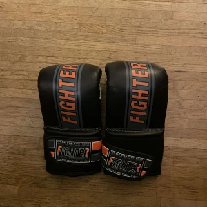  boxningshandskar från Fighter - Snygga svarta boxningshandskar från Fighter med orange och grå detaljer. Perfekta för träning, de har en robust design och breda kardborreband för extra stöd. Handskarna har en ventilerad insida för bättre komfort under intensiva pass.
