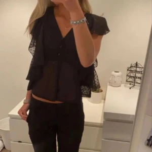 Topp från bikbok - Snygg svart blus med volangdetaljer och knappar framtill. Blusen har korta ärmar och en luftig passform, perfekt för en stilren look.