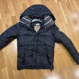 Svart pufferjacka från Burberry. S-M - Säljer en snygg svart pufferjacka från Burberry med klassiskt rutigt foder. Jackan har dragkedja och knappar framtill samt två praktiska fickor med dragkedja. Perfekt för kyliga dagar! Storlek S-M