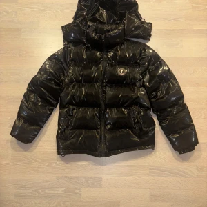 Svart pufferjacka (Trapstar)  - Snygg svart pufferjacka med glansig finish och huva. Jackan har en dragkedja framtill och ett märke på bröstet. Perfekt för kyliga dagar när du vill hålla stilen. Ops ändå negativa är att dragkedjan till luvan går inte att öppna. //, Elliot