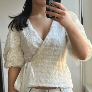 Vit omlottblus med knytband - Säljer en söt vit omlottblus med knytband i midjan. Blusen har korta puffärmar och ett fint texturerat mönster. Perfekt för en somrig look! Nästan oanvänd!💗