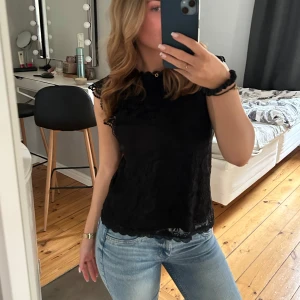 Svart spetsblus - Säljer en elegant svart spetsblus med fina spetsdetaljer vid ärmarna. Blusen är ärmlös och har en rund halsringning. Perfekt för att ge en stilren look.