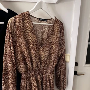 Brun mönstrad klänning från Zara - Snygg brun klänning från Zara med ett djurinspirerat mönster. Klänningen har en v-ringad hals och långa ärmar, perfekt för en trendig look. Den elastiska midjan ger en smickrande passform. Perfekt för att sticka ut i mängden! Fler bilder går att få. M men skulle säga strl S mer.