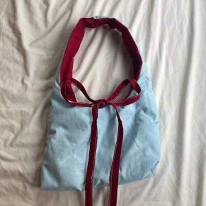 Blå axelväska med röda detaljer - Handgjord! Snygg blå axelväska med kontrasterande röda detaljer och knytband. Perfekt för att ge en färgklick till din outfit. Väskan har en enkel och stilren design som passar till många tillfällen.